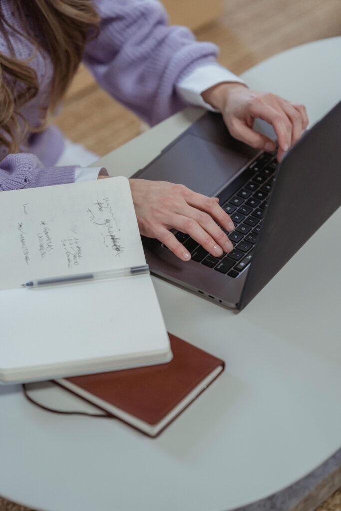 From above of unrecognizable female worker browsing internet on netbook while sitting at desk with opened notebook in light room